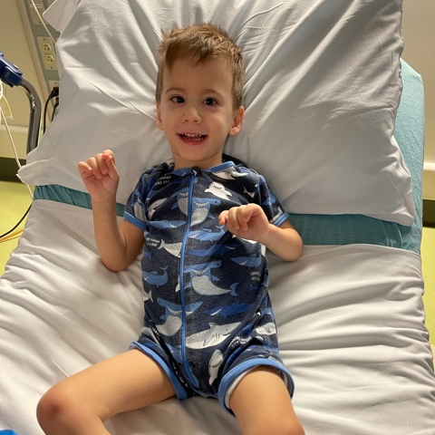 a young boy in a hospital bed. He is wearing a blue romper with whales and is looking at the camera smiling