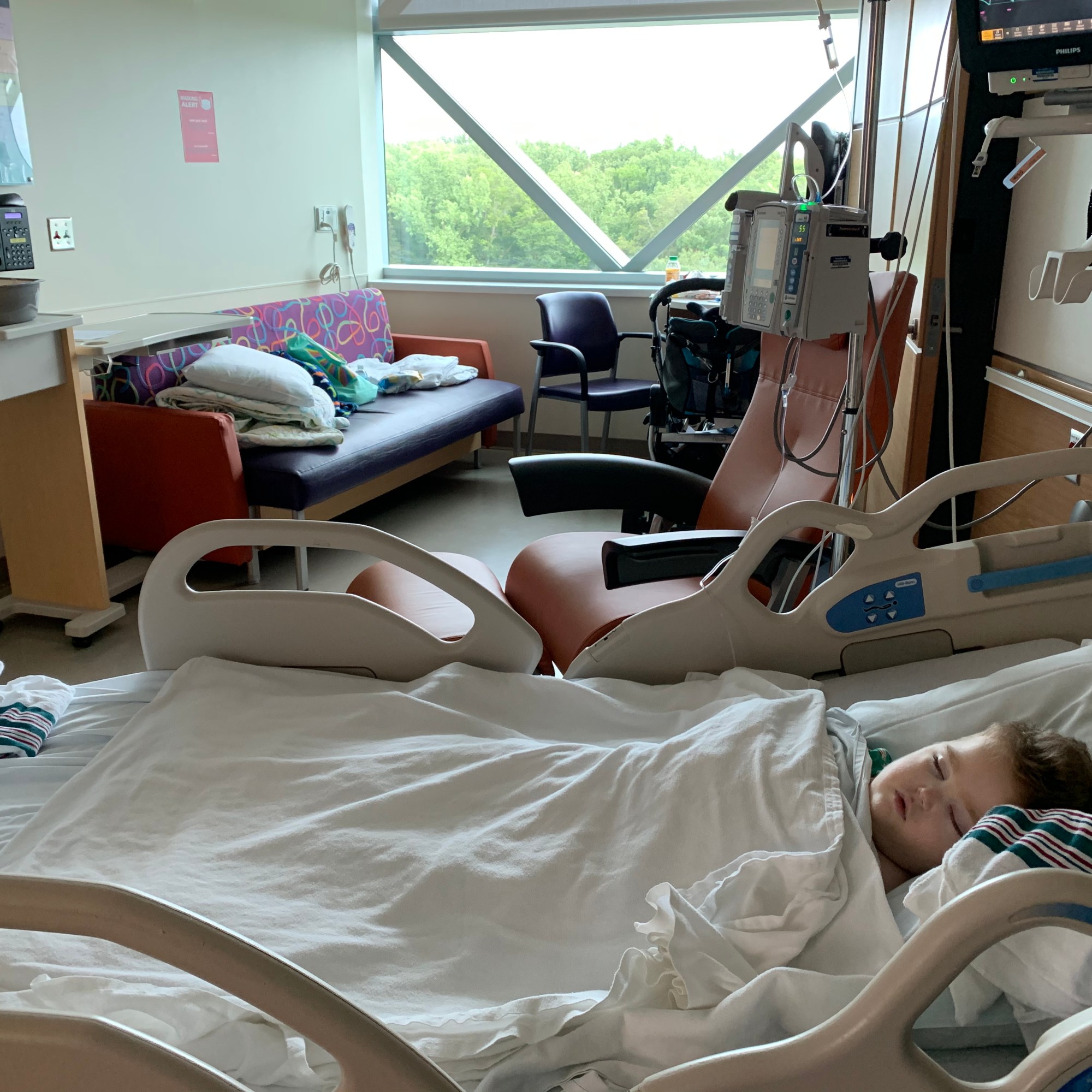 a 3 year old boy sleeping in a hospital bed. The room is lit by natural light and has a window that overlooks trees.