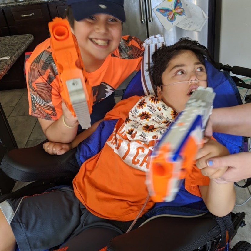 two boys in bright orange shirts holding Nerf guns. One boy is in a wheelchair and is receiving some support to hold the Nerf gun. He has nasal cannulas for oxygen support. The other boy is leaning on the wheelchair looking at the camera. Both boys have big smiles.
