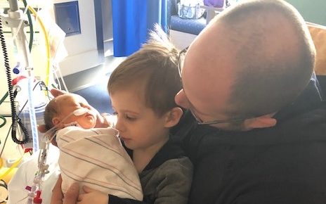 A boy about 3 years old is sitting on his father's lap while holding his newborn baby brother. They are in a NICU hospital room. The baby is wrapped in the blanket and has oxygen support.
