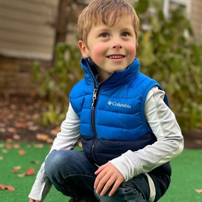 Boy with brown hair smiling and kneeling outside on grass wearing a blue vest and jeans
