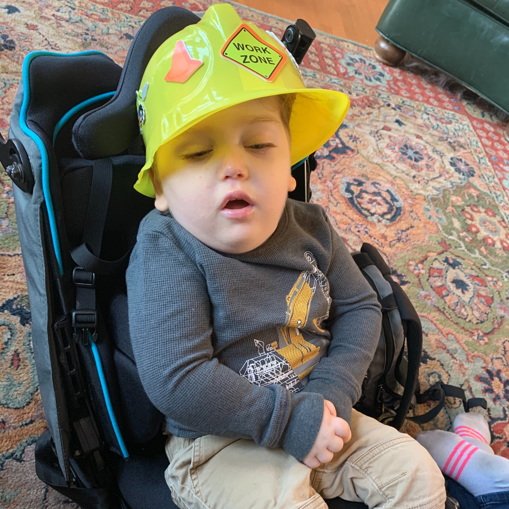 A boy is wearing a plastic construction hat at a child's birthday party. He is wearing a shirt with a construction vehicle on it and is sitting in a wheelchair stroller.