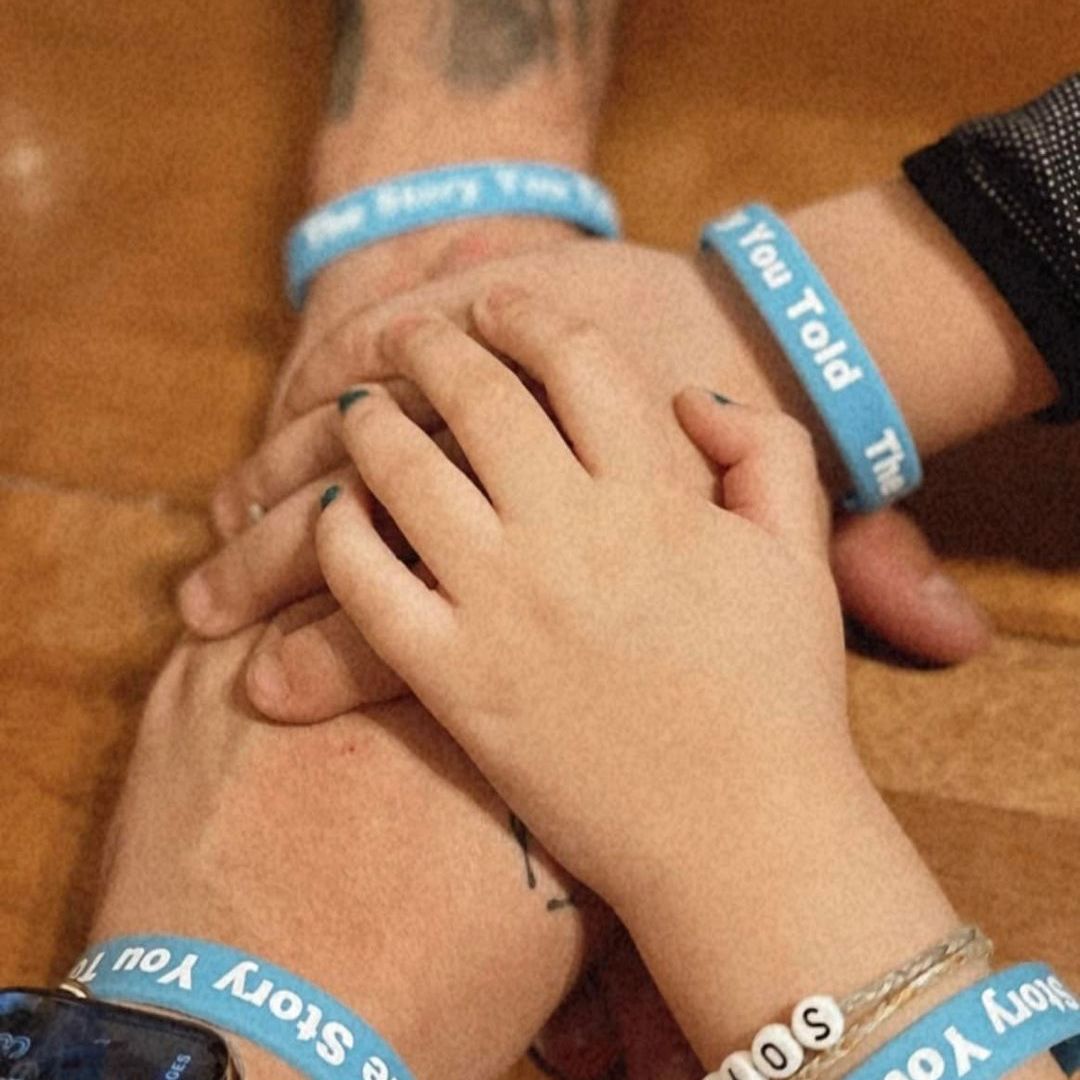 two adult and two child hands piled on each other. All four of them are wearing blue The Story You Told wrist bands