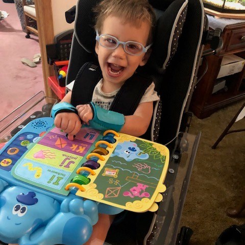 A young boy with blue glasses sitting in a supported seat. The seat has a tray and a Blue's Blue's book is open on the tray. He has on wrist braces and a very big smile.