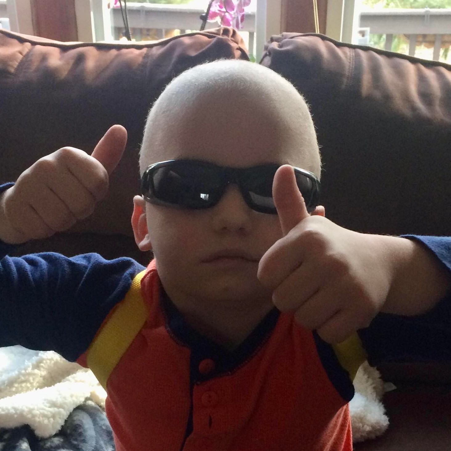 Dominic is sitting on a couch wearing sporty sunglasses. He is holding two thumbs up.