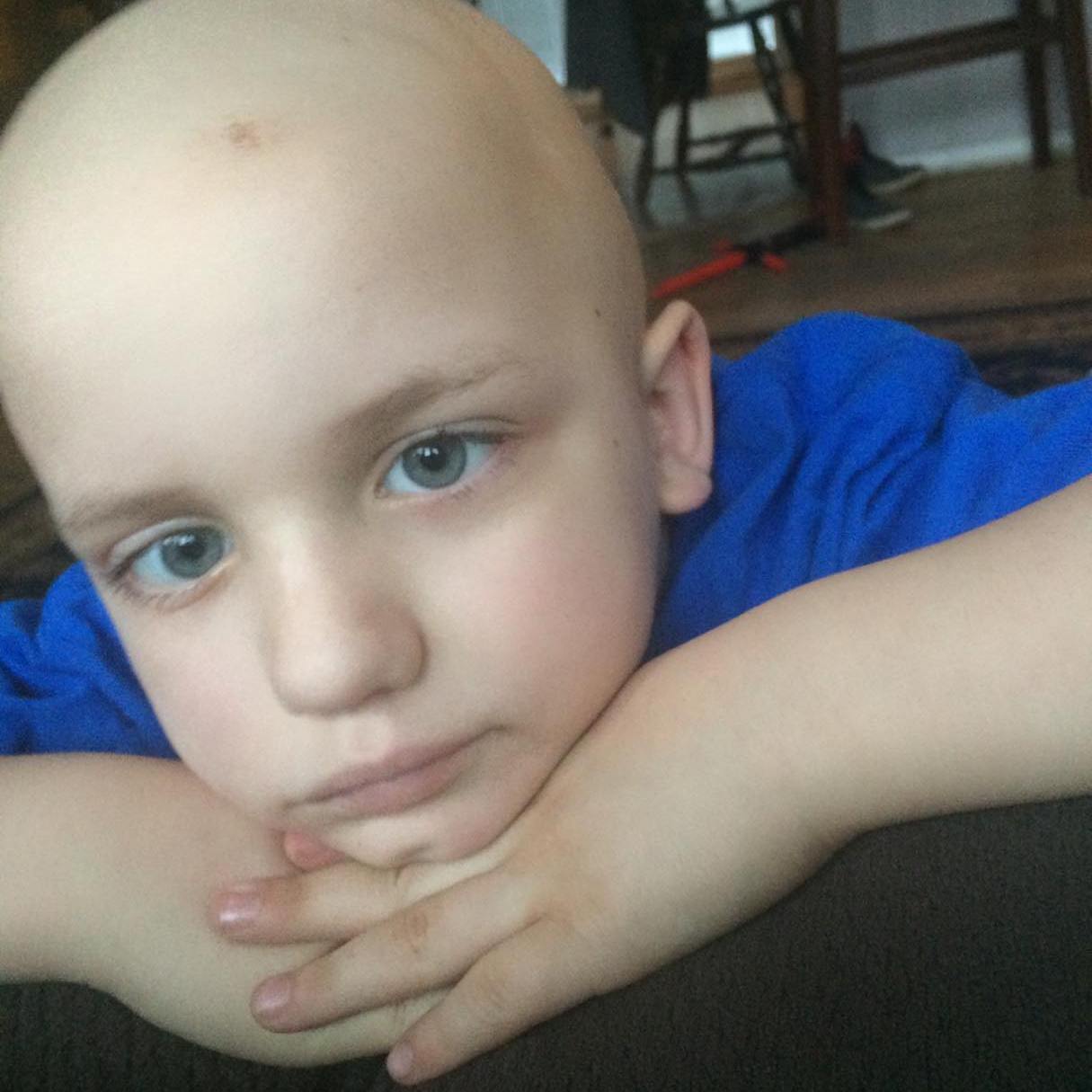 Dominic leans against the back of a couch. His face is resting on the back of his hands and he has a subtle smile. He has bright blue eyes and is wearing a blue shirt.